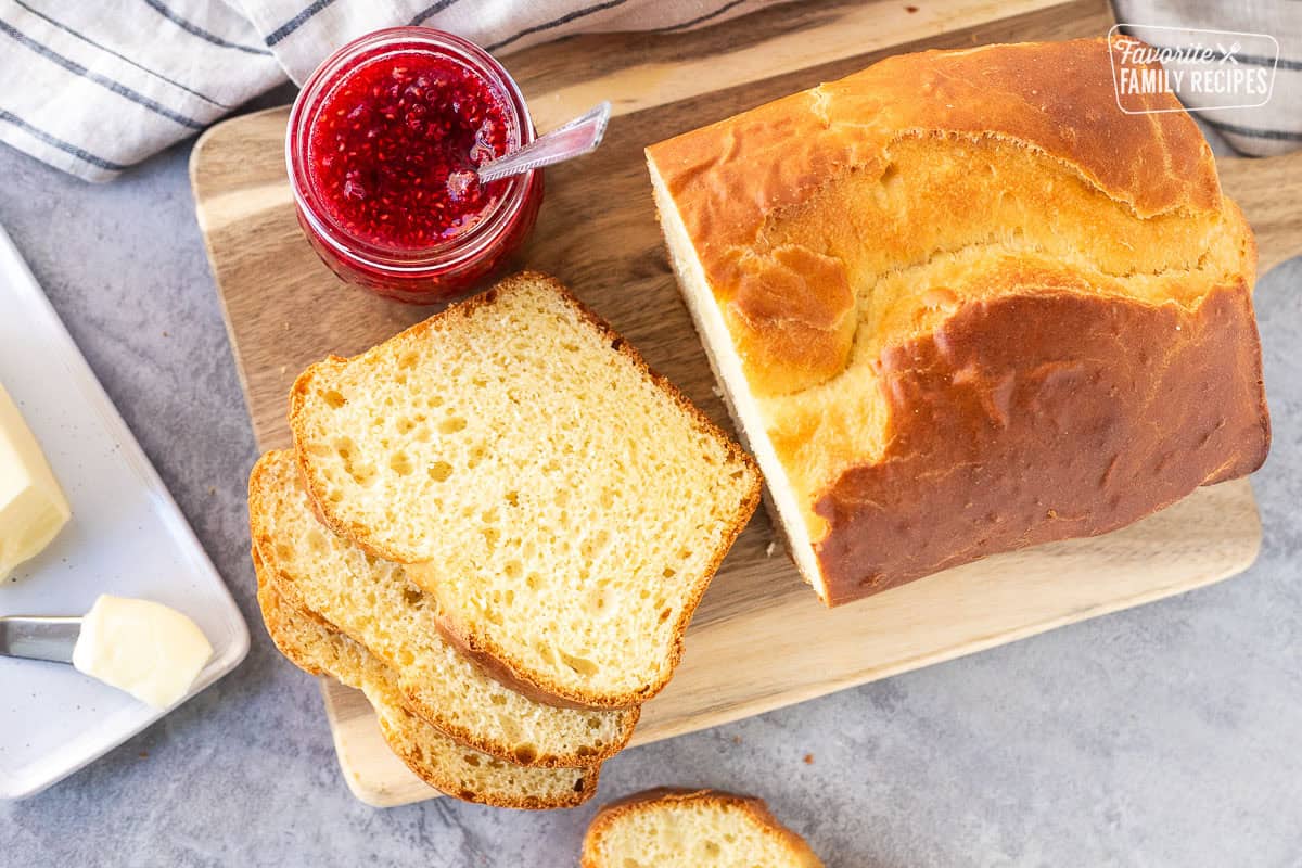 This Sally Lunn bread recipe comes to us from my great-grandmother. This sweet bread is a family favorite, passed down through generations and is always warm, sweet and satisfying. Sally Lunn Bread sliced on wooden board. Butter dish and raspberry jam on the side.