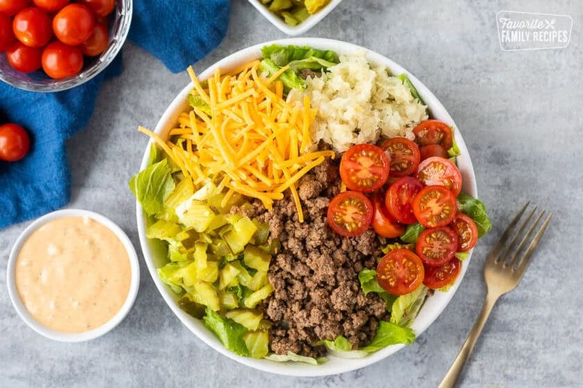 Burger Salad in a bowl with lettuce, hamburger meat, cherry tomatoes, grilled onions, cheddar cheese, chopped dill pickles and special sauce dressing on the side.