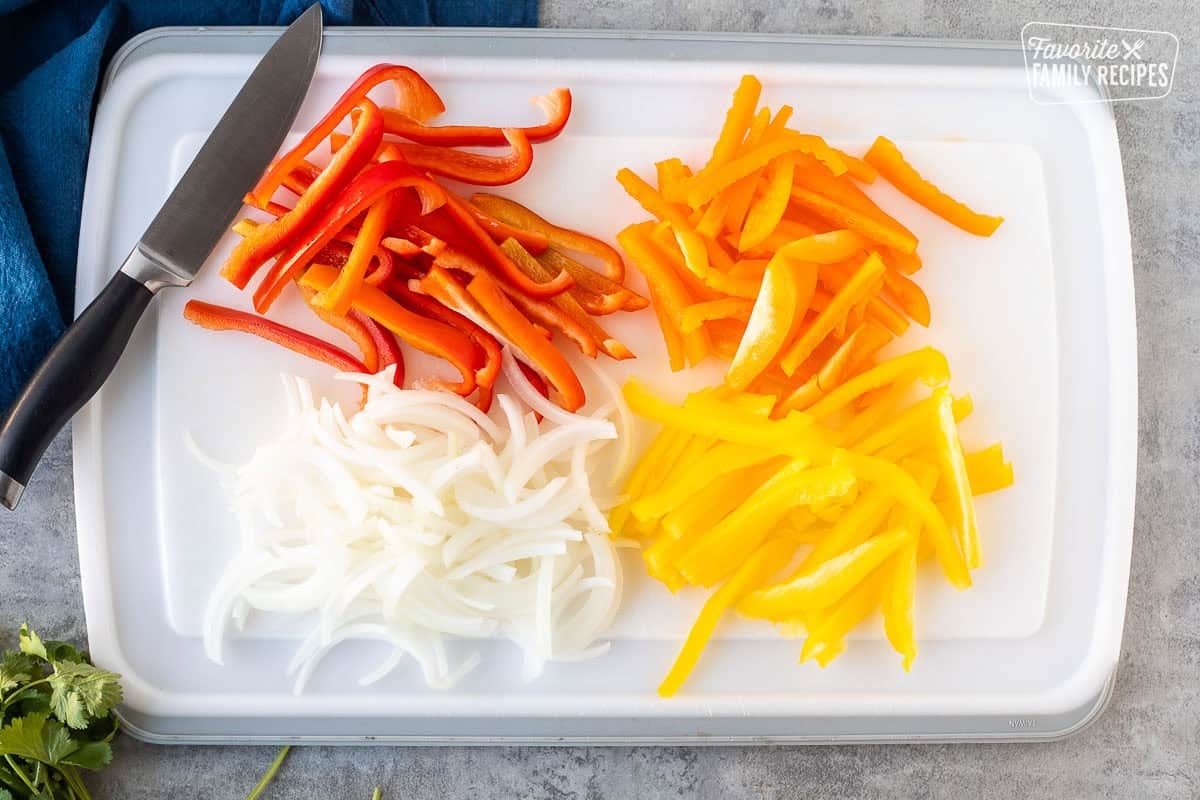 Cutting board with cut red bell peppers, orange bell peppers, yellow bell peppers and onion with knife.
