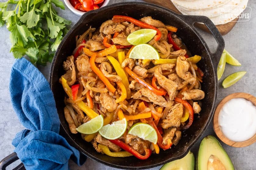 Skillet for chicken fajitas with marinated chicken, onion and bell peppers. Sides of flour tortillas, avocado, sour cream limes, cilantro and tomatoes.