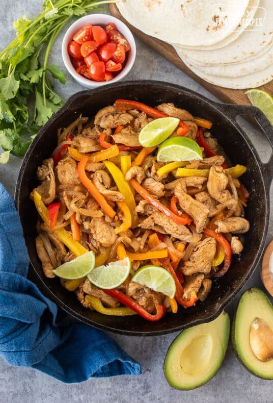 Skillet for chicken fajitas with marinated chicken, onion and bell peppers. Sides of flour tortillas, avocado, sour cream limes, cilantro and tomatoes.