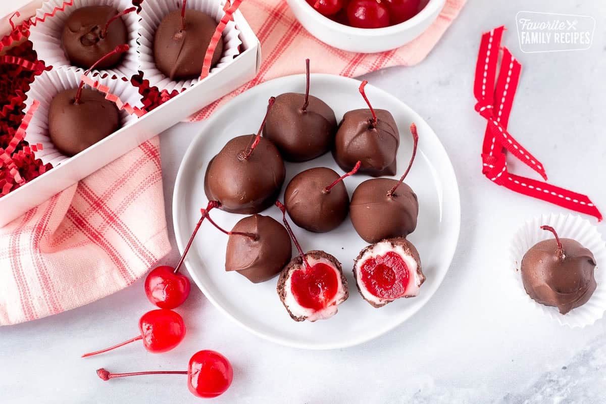 Plate with chocolate covered cherries with one cut open in half next to box of cherries.
