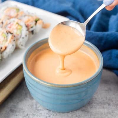 Drizzling easy sriracha mayo from a spoon into a bowl. Plate of sushi on the side.