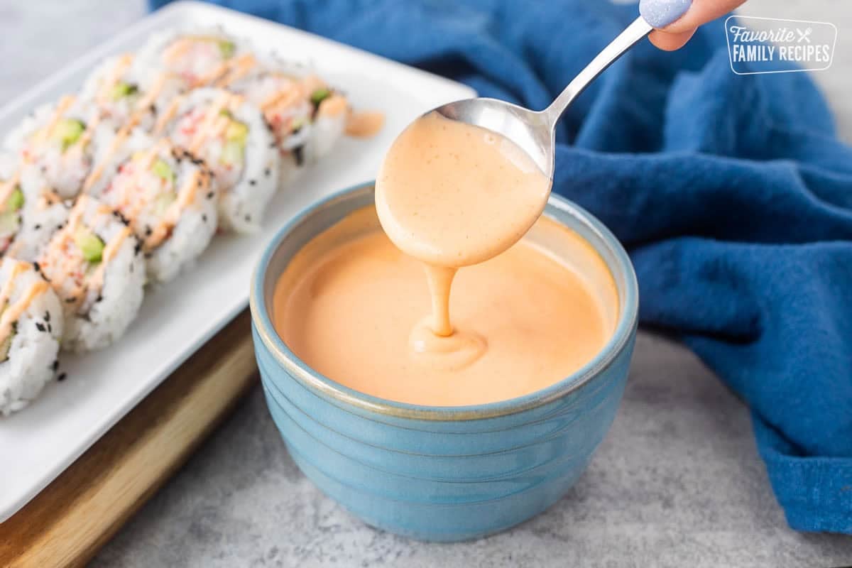 Drizzling easy sriracha mayo from a spoon into a bowl. Plate of sushi on the side.
