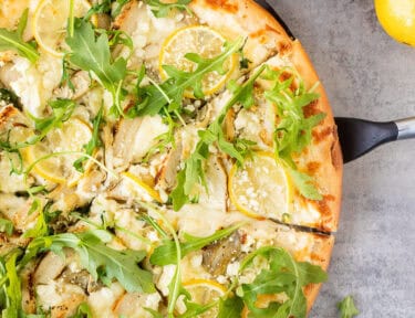 Spatula under slice of lemon chicken pizza with cheesy lemon white sauce, mozzarella cheese, artichoke hearts, lemon chicken, lemon slices, feta cheese and fresh arugula.