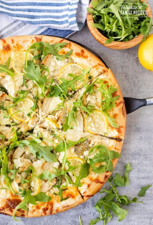 Spatula under slice of lemon chicken pizza with cheesy lemon white sauce, mozzarella cheese, artichoke hearts, lemon chicken, lemon slices, feta cheese and fresh arugula.