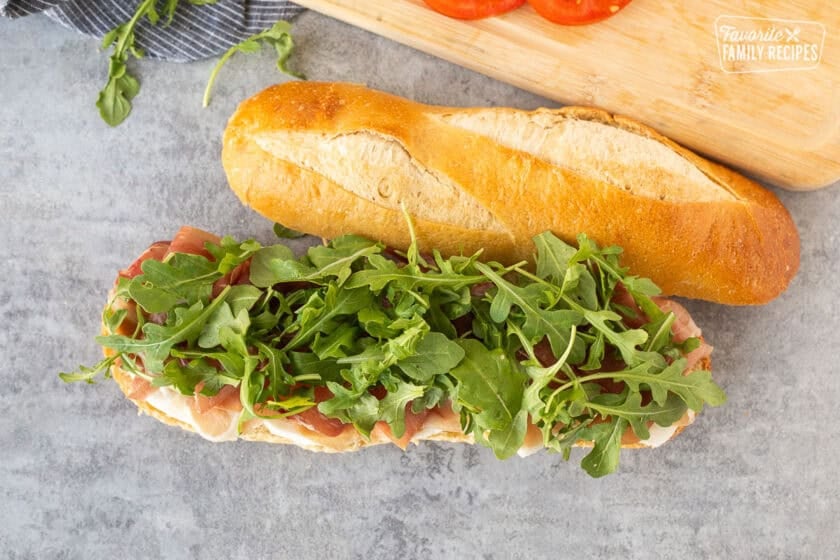 Demi French bread cut in half with a layer of fresh thick cut mozzarella cheese, prosciutto and arugula.
