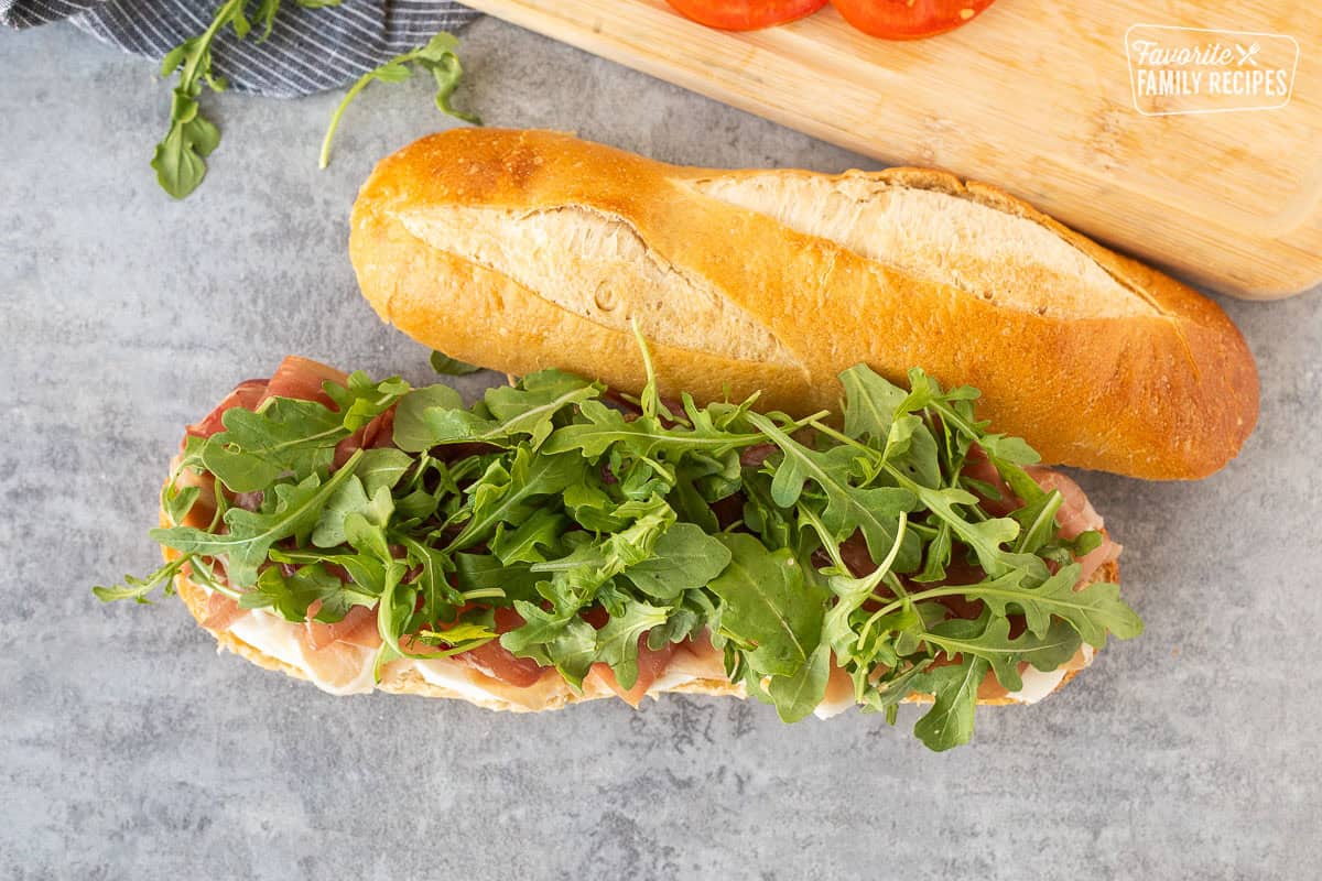 Demi French bread cut in half with a layer of fresh thick cut mozzarella cheese, prosciutto and arugula.