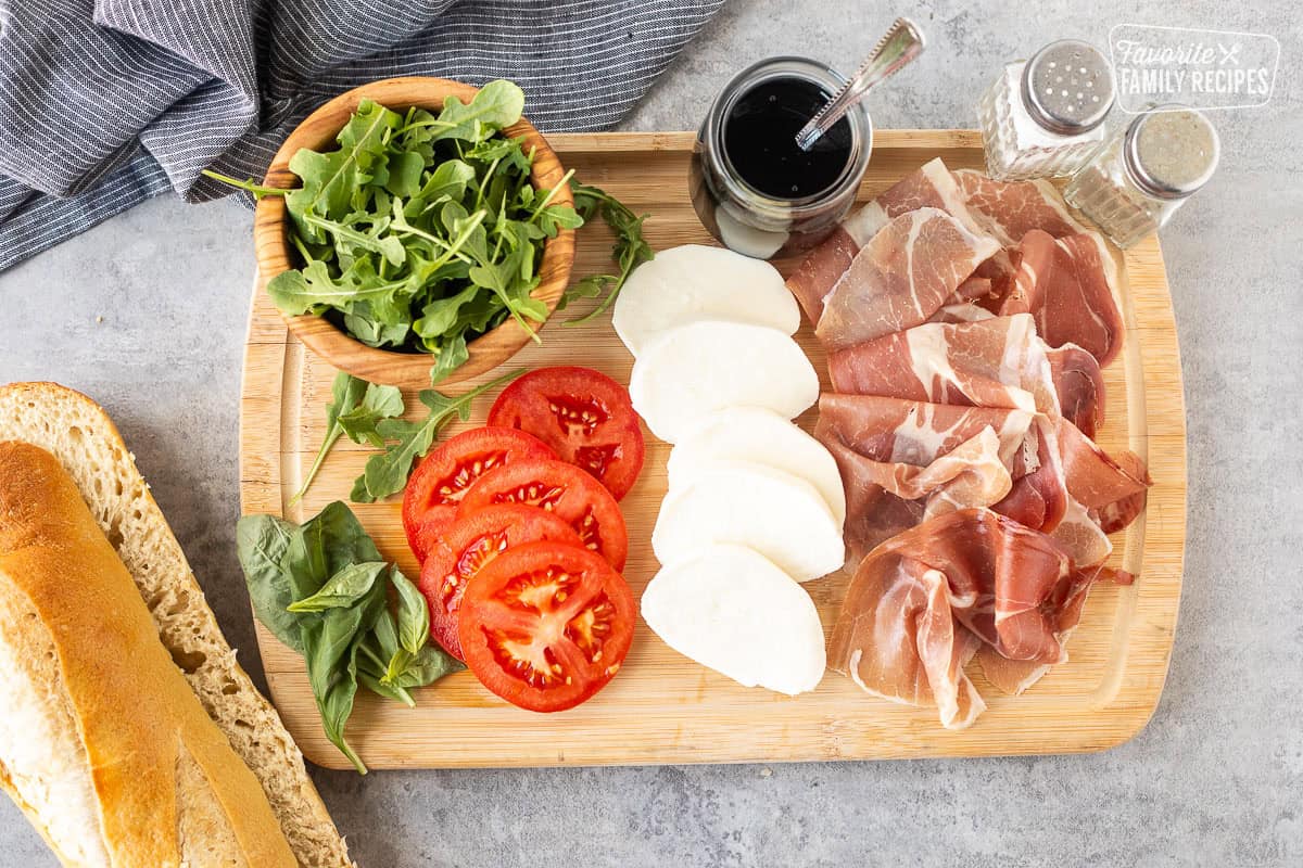 Cutting board with balsamic glaze, mozzarella cheese, prosciutto, basil, arugula, tomato, salt and pepper. Demi French bread on the side cut.