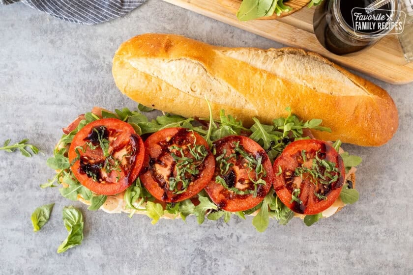 Demi French bread cut in half with a layer of fresh thick cut mozzarella cheese, prosciutto, arugula, tomatoes, basil and balsamic glaze.