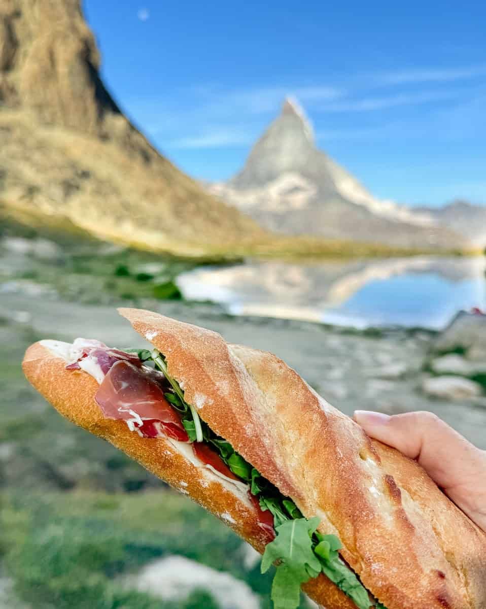 A prosciutto sandwich being held up in front of a lake during a hike with the Matterhorn in Switzerland in the background.