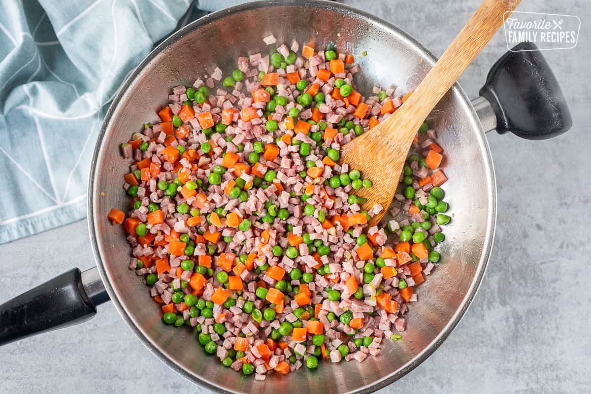 Skillet of ham, peas and carrots with a wooden spoon.