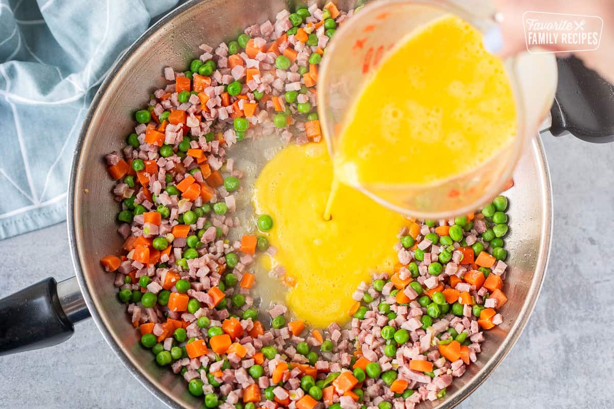 Pouring egg into skillet of ham, peas and carrots.