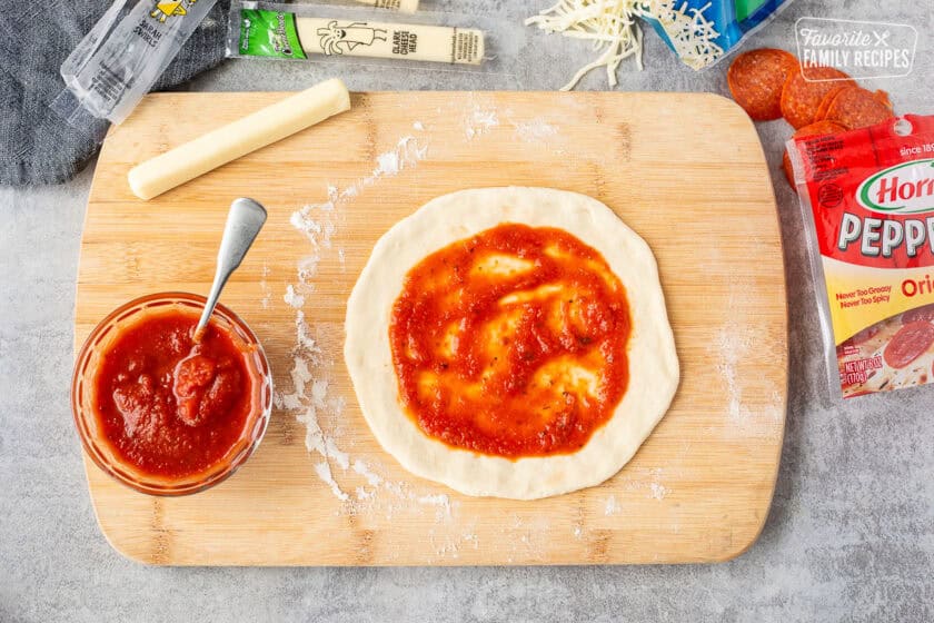 Spreading sauce on pizza dough on a cutting board.