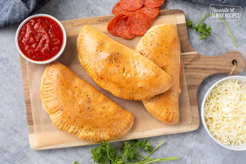 Three baked easy calzone recipe next to small bowl of marinara sauce, mozzarella cheese and pepperoni.