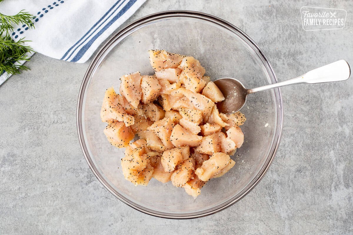 Bowl of cut chicken tossed in lemon pepper seasoning, salt and pepper with spoon.