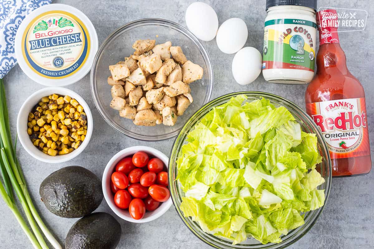 Ingredients to make buffalo chicken salad including chopped lettuce, tomatoes, diced chicken, fire roasted corn, avocado, blue cheese, green onion, hard boiled eggs, ranch dressing and Frank's red hot sauce.