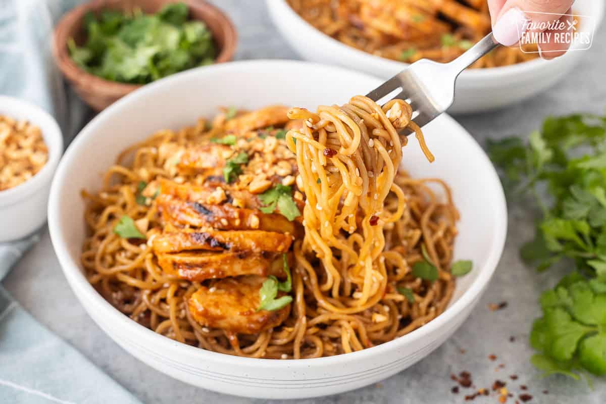 Fork holding sweet and spicy noodles from a bowl with grilled chicken.