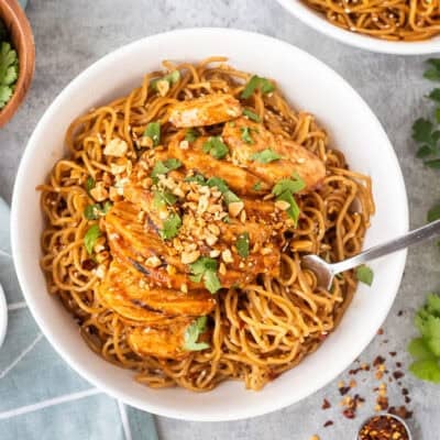 Sweet and spicy noodles with grilled chicken in a bowl with a fork and topped with cilantro and peanuts.