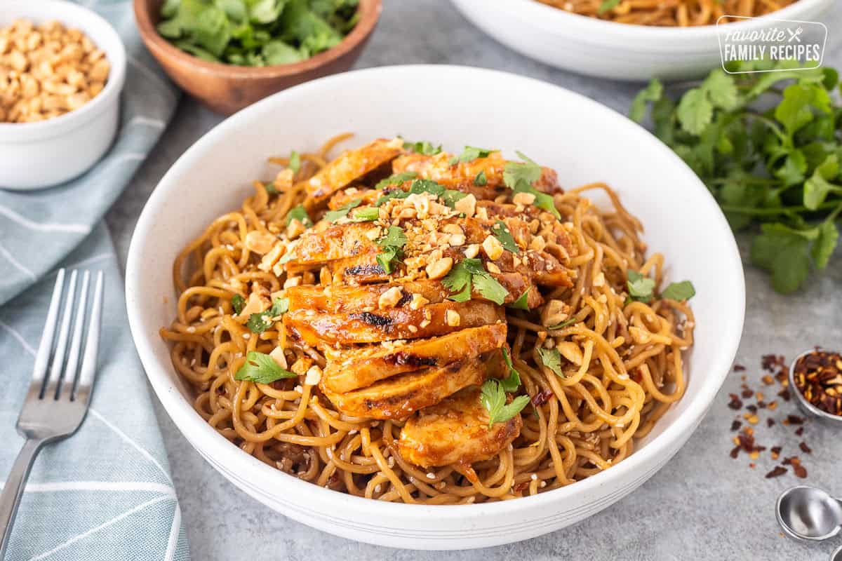 Sweet and spicy noodles with grilled chicken in a bowl with a fork and topped with cilantro and peanuts.