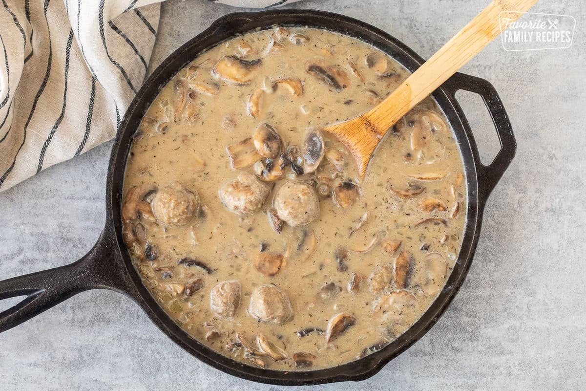 Meatballs added to creamy stroganoff sauce in a skillet with wooden spoon.