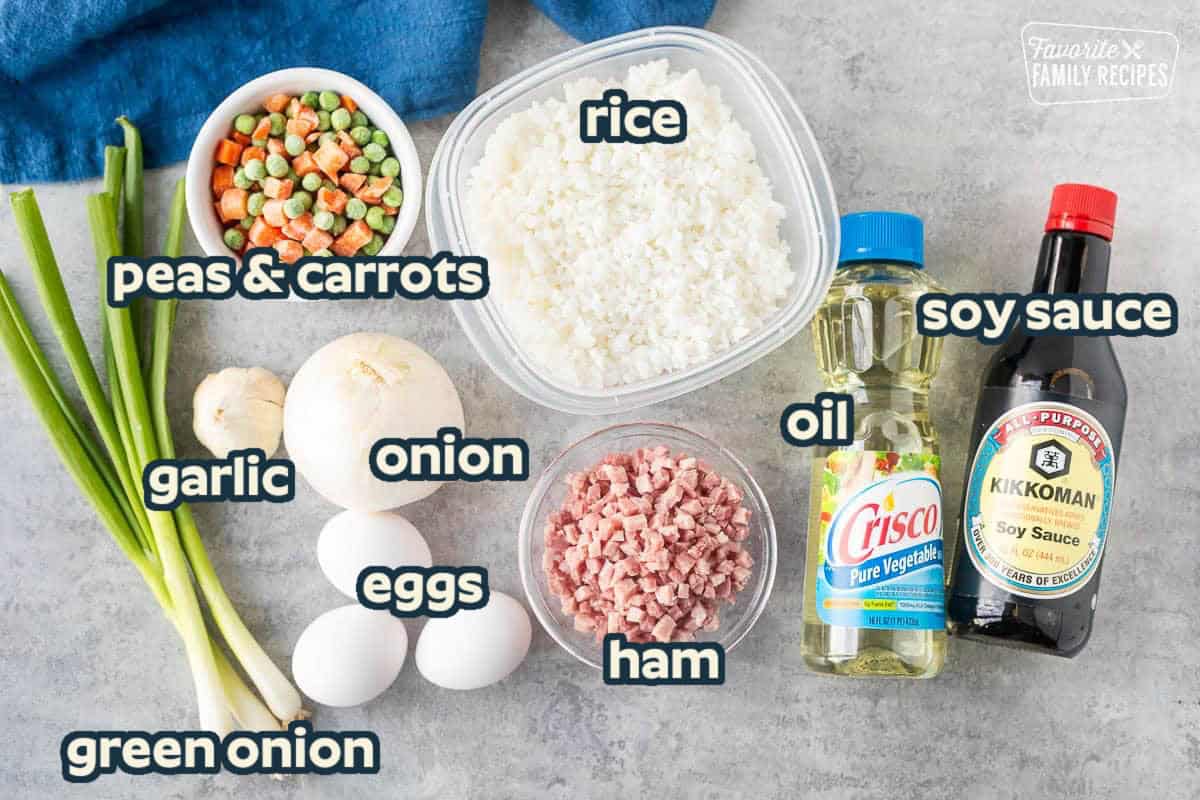 Ingredients to make Ham Fried Rice including day old rice, soy sauce, vegetable oil, small pieces of ham, eggs, onion, garlic, frozen carrots and peas. Green onion for garnish.