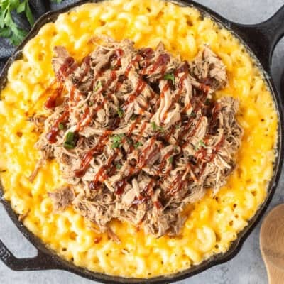 Baked macaroni and cheese in a skillet with pulled pork over the top and drizzled with barbecue sauce.