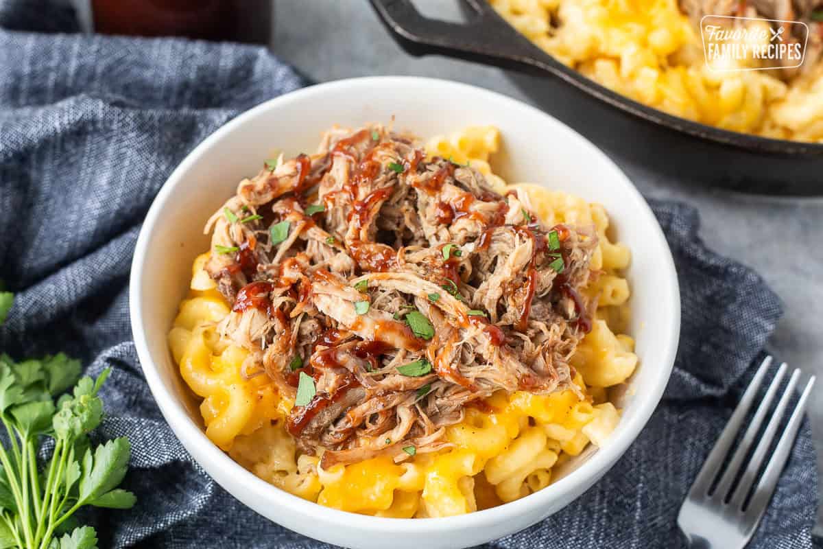 A bowl of macaroni and cheese with pulled pork over the top and drizzled with barbecue sauce next to a cast iron skillet.