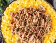 Baked macaroni and cheese in a skillet with pulled pork over the top and drizzled with barbecue sauce.