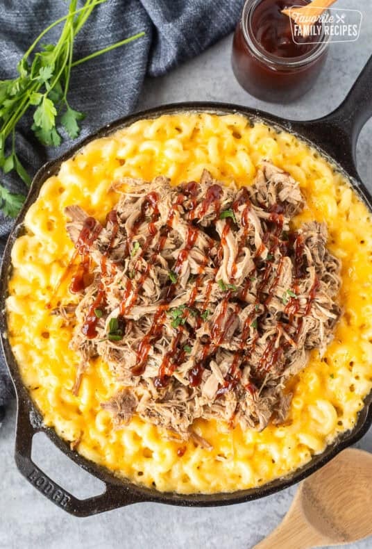 Baked macaroni and cheese in a skillet with pulled pork over the top and drizzled with barbecue sauce.