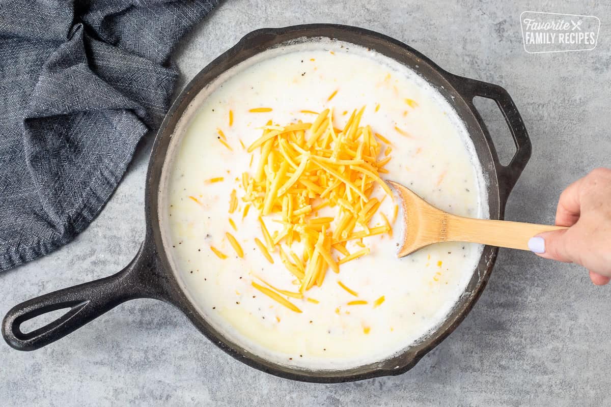 A cast iron skillet with cream sauce with shredded yellow cheese over the top that hasn't been incorporated yet into the sauce to make mac and cheese