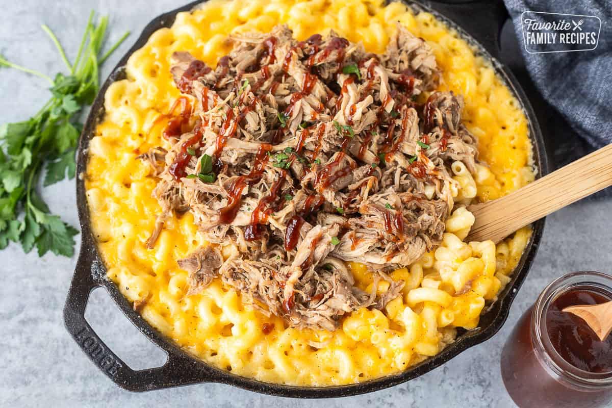 Baked macaroni and cheese in a skillet with pulled pork over the top and drizzled with barbecue sauce. A wooden spoon is placed in the skillet to serve the macaroni and cheese.