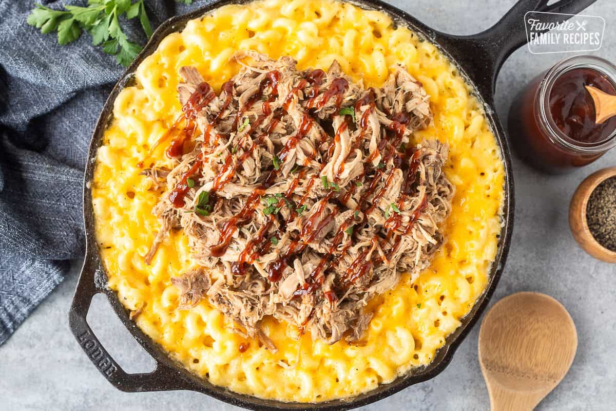 Baked macaroni and cheese in a skillet with pulled pork over the top and drizzled with barbecue sauce.