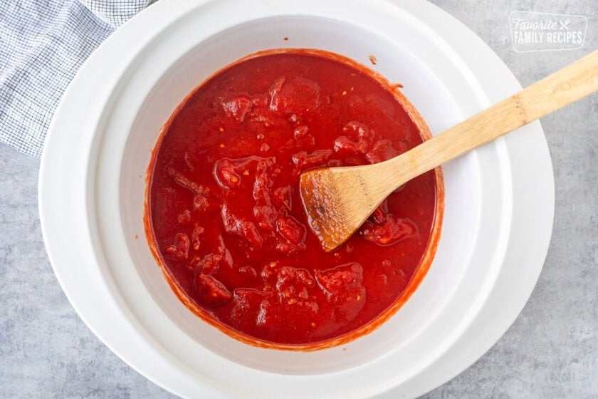 Crockpot of broken up canned tomatoes with a wooden spoon.