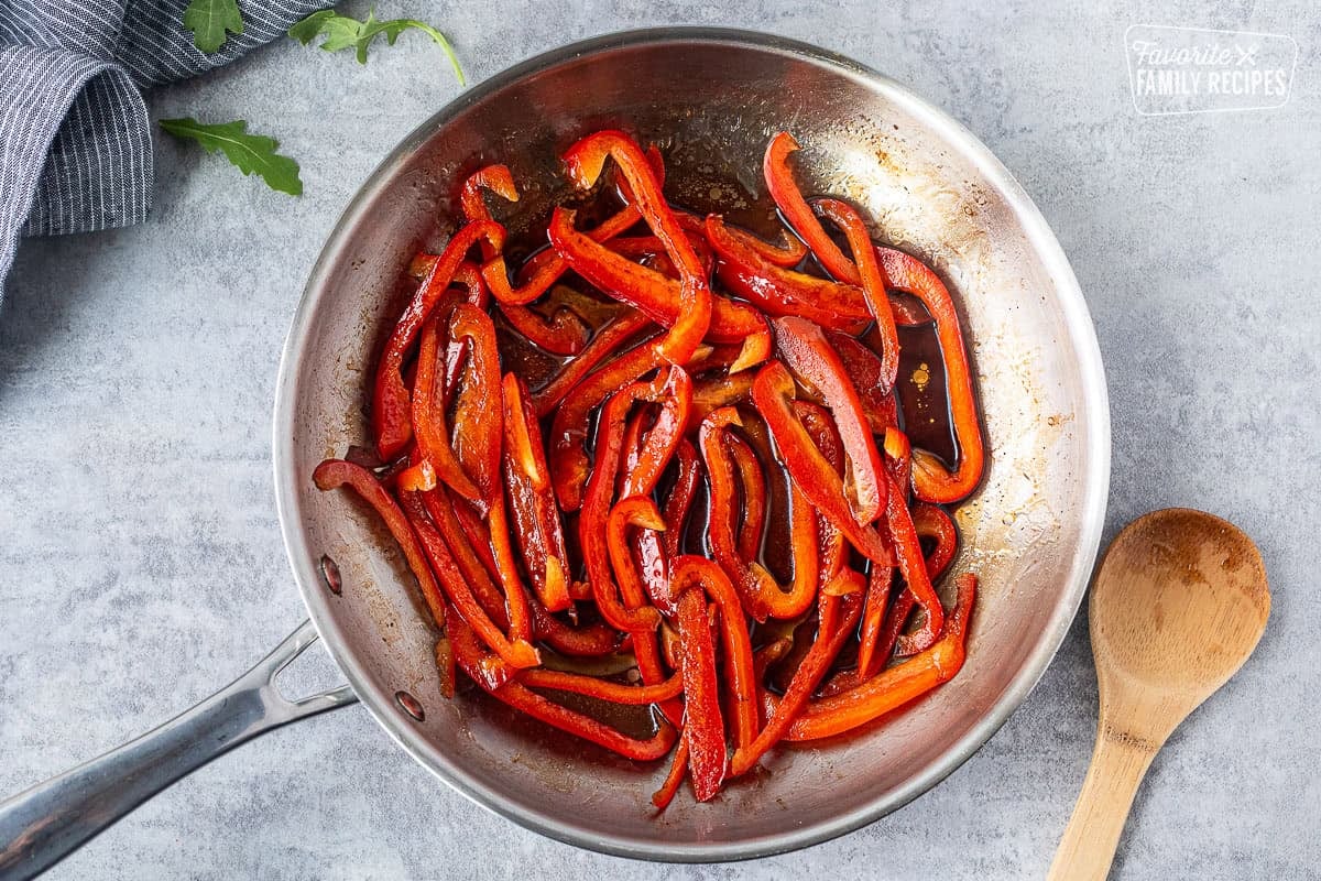 Skillet with cut red peppers and balsamic vinegar with wooden spoon on the side.
