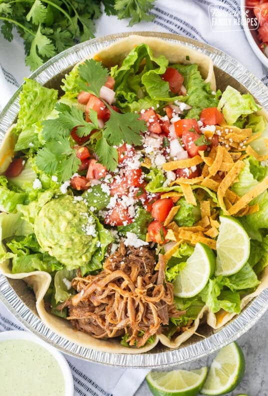 Cafe Rio Sweet Pork salad in a metal dish with lime wedges, pico de Gallo, cilantro, guacamole, cojita cheese and side of cilantro ranch dressing.