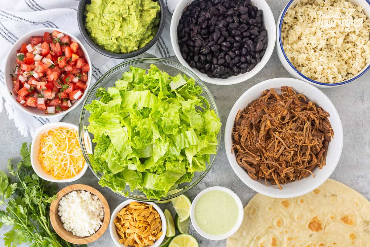 Ingredients to make Cafe Rio Sweet Pork Salad including lettuce, sweet pork, black beans, cilantro lime rice, guacamole, pico de Gallo, shredded Mexican blend cheese, cojita cheese, tortilla strips, lime, tortilla and cilantro ranch dressing.