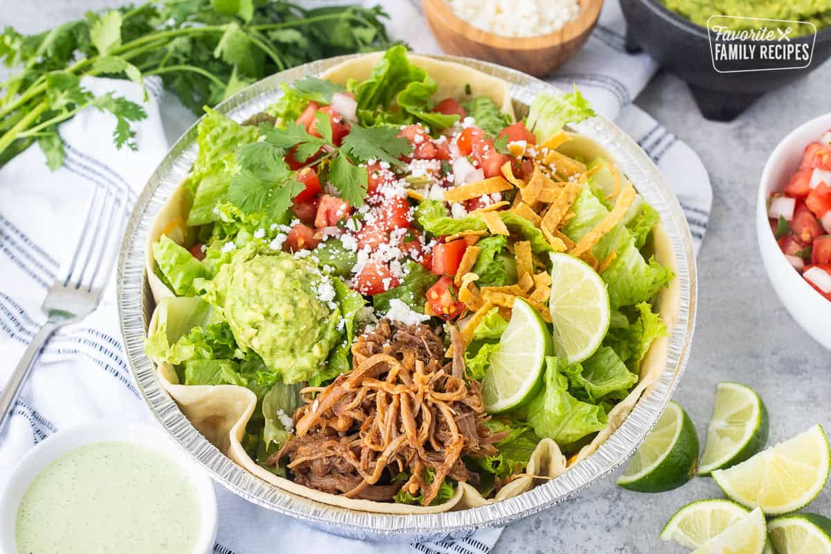 Cafe Rio Sweet Pork salad in a metal dish with lime wedges, pico de Gallo, cilantro, guacamole, cojita cheese and side of cilantro ranch dressing.