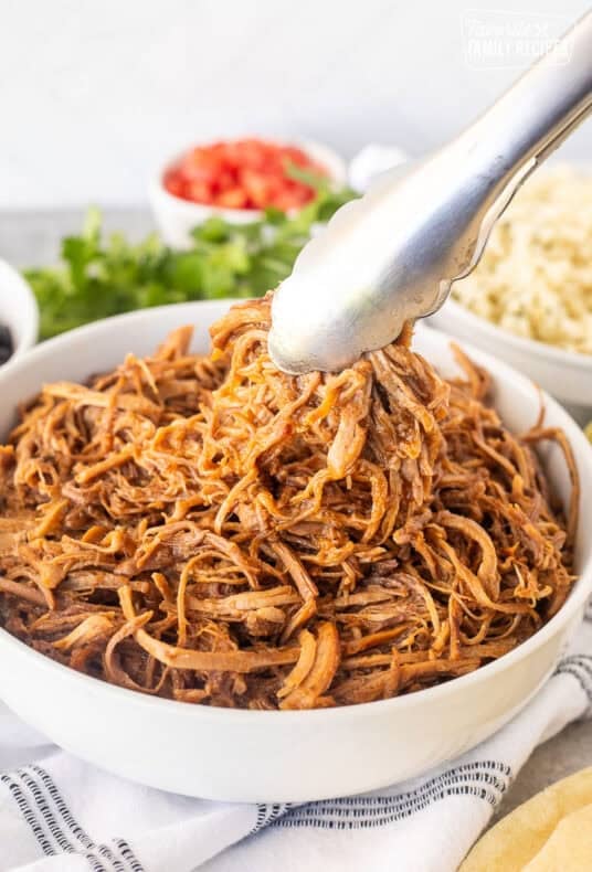 Bowl of Cafe Rio Sweet Pork with tongs. Sides of rice and black beans on the side.