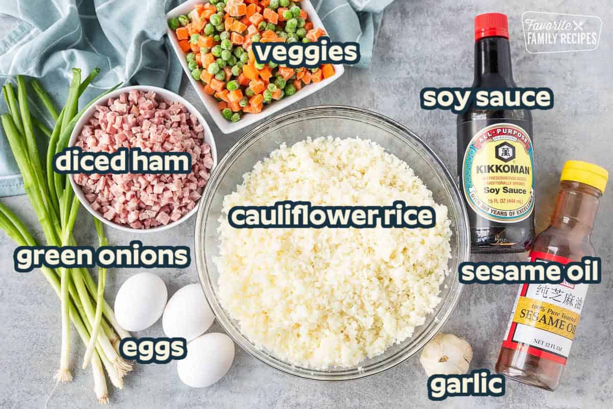 Ingredients to make cauliflower fried rice including cauliflower rice, soy sauce, sesame oil, garlic, egg, green onion, ham, and frozen peas and carrots.