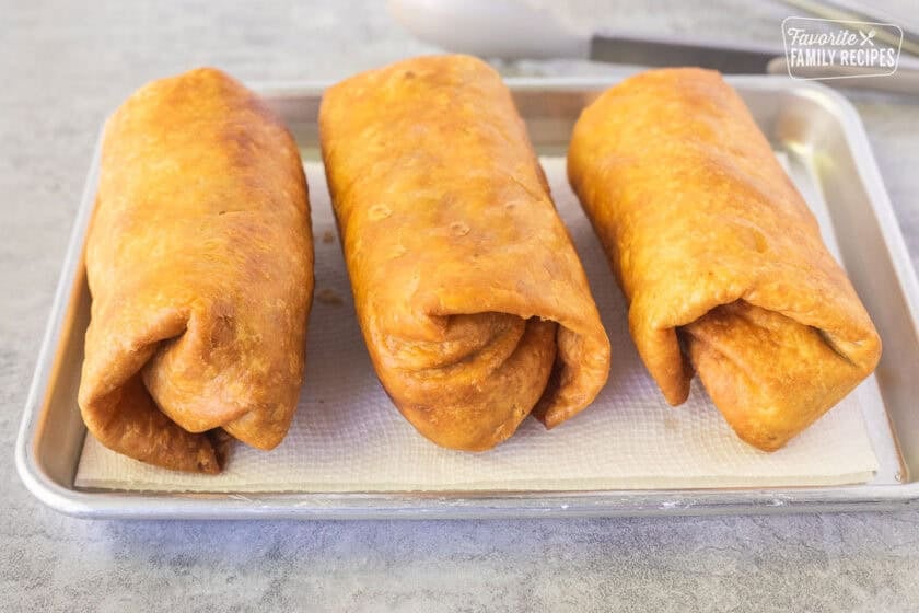 Three fried Chicken Chimichangas on a paper towel on a baking dish.