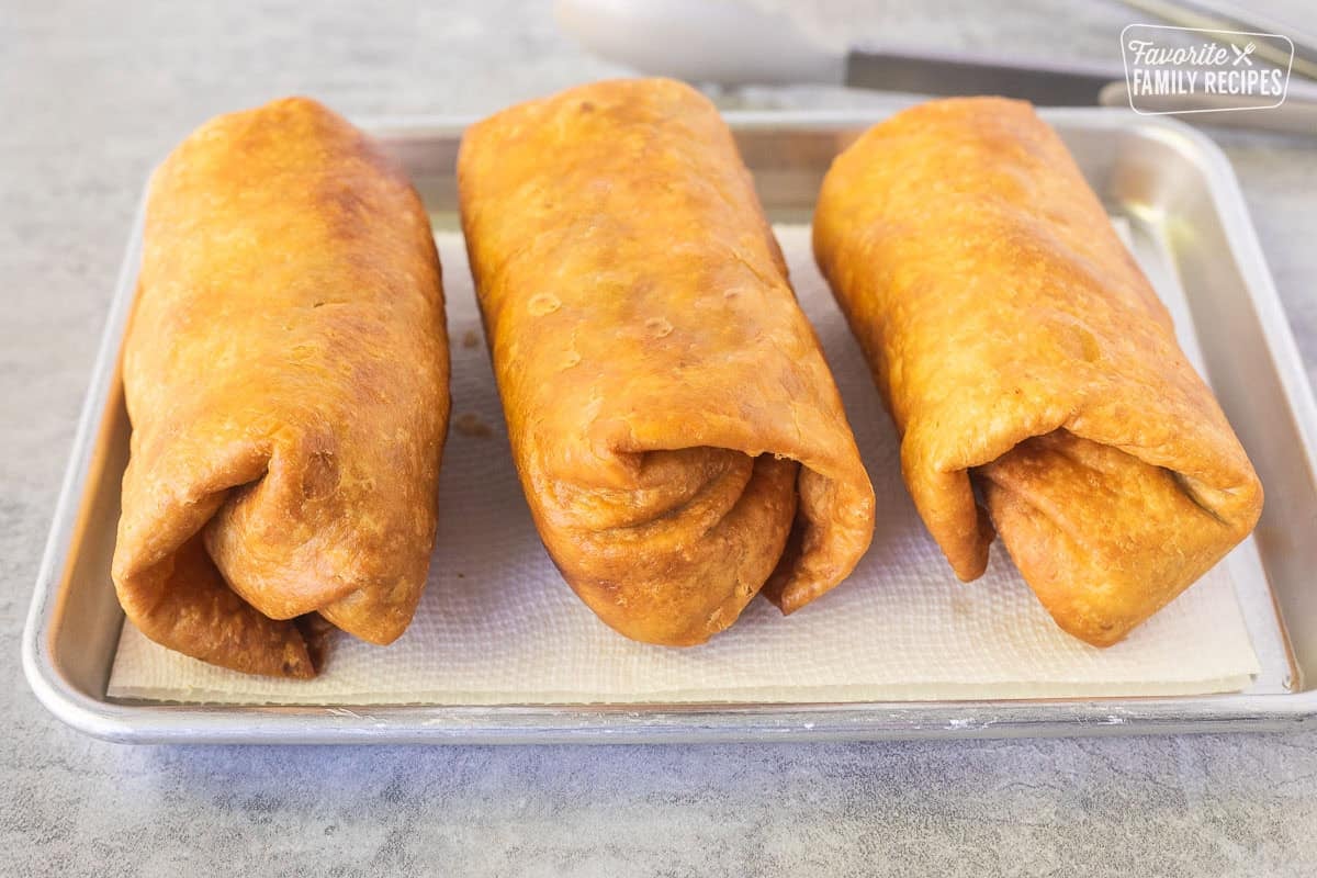 Three fried Chicken Chimichangas on a paper towel on a baking dish.
