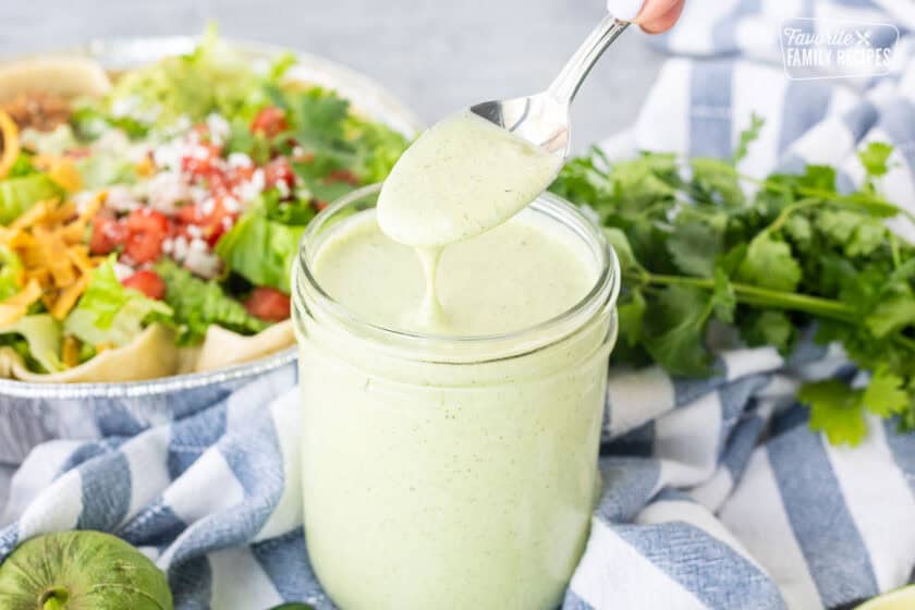Spoon drizzling Cafe Rio Creamy Tomatillo Ranch Dressing into jar with salad in the background.