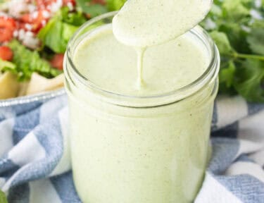 Spoon drizzling Cafe Rio Creamy Tomatillo Ranch Dressing into jar with salad in the background.