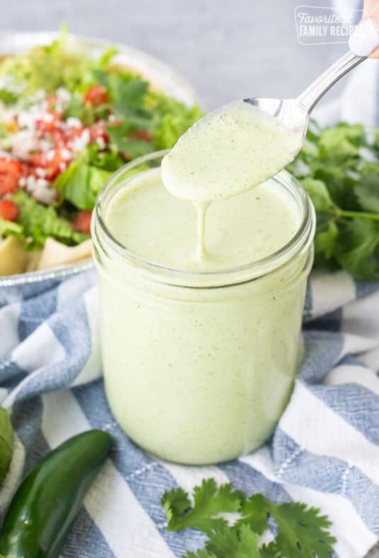 Spoon drizzling Cafe Rio Creamy Tomatillo Ranch Dressing into jar with salad in the background.