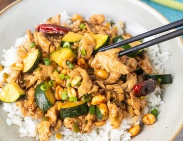 Chop sticks picking up a piece of kung pao chicken from bowl over rice.