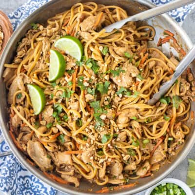 Skillet of Thai peanut noodles topped with chopped peanuts, lime wedges and chopped cilantro.