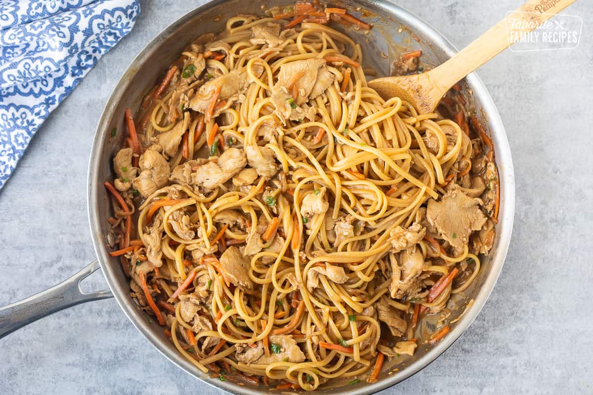 Noodles added to skillet of peanut chicken sauce and wooden spoon.