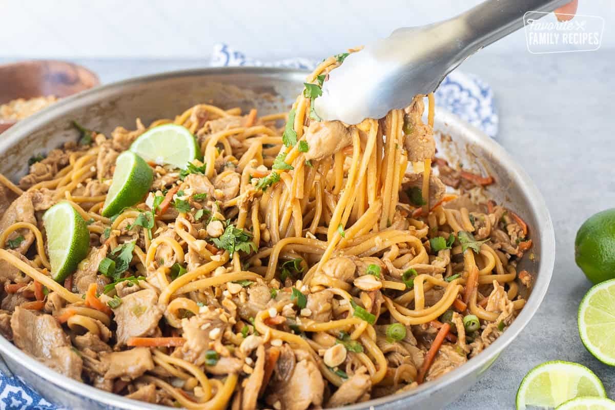 Tongs with Thai peanut chicken noodles topped with chopped peanuts, lime wedges and chopped cilantro.