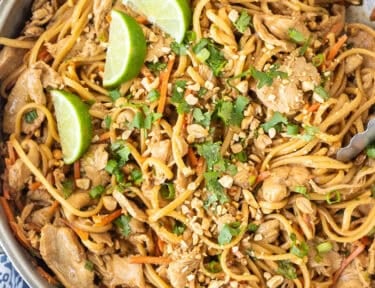 Skillet of Thai peanut noodles topped with chopped peanuts, lime wedges and chopped cilantro.